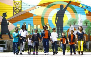 8 Charlotte kids in a line holding hands walking towards you with a beautiful mural on a building in the background.