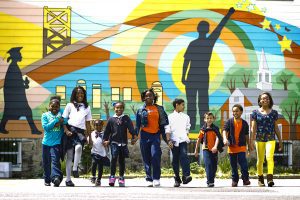 8 Charlotte kids in a line holding hands walking towards you with a beautiful mural on a building in the background.