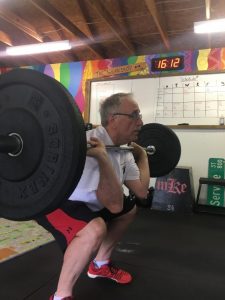 A man squatting a barbell in a colorful gym. He's wearing a white tshirt and red and black shorts.