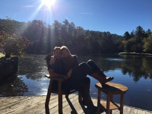 Meet Jan Tiffany. Jan is sitting happily on her husbands lap in a wood chair in front of a lake with the sun beaming behind.