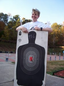 Retired CMPD Officer Becomes Fitness Coach, lady with short light brown hair in front of a shooting target that says 99.6%