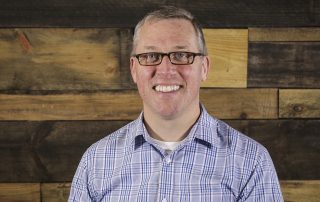Three Charlotte Pastors Win Elite Wellness Package MissionFiT, headshot, man in his 40's, standing in front of a wood palat wall with a blue dress shirt on and glasses
