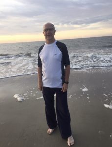 Today's Spotlight Athlete, Pastor Brent Watts, pastor standing that the brim of the ocean with a beautiful sky and water behing him, he is wearing black glasses and and black and white t-shirt