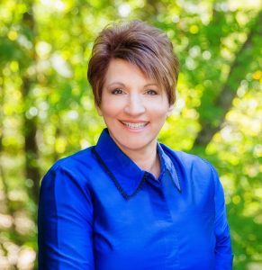 Testimony Tuesday - National Speaker, Zoe Elmore, Zoe's headshot, she is sitting smiling with a bright blue dress shirt and green trees in the background