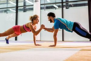 Husband and wife in a plank posistion in a gym clasping one anothers fists symbolizing a team