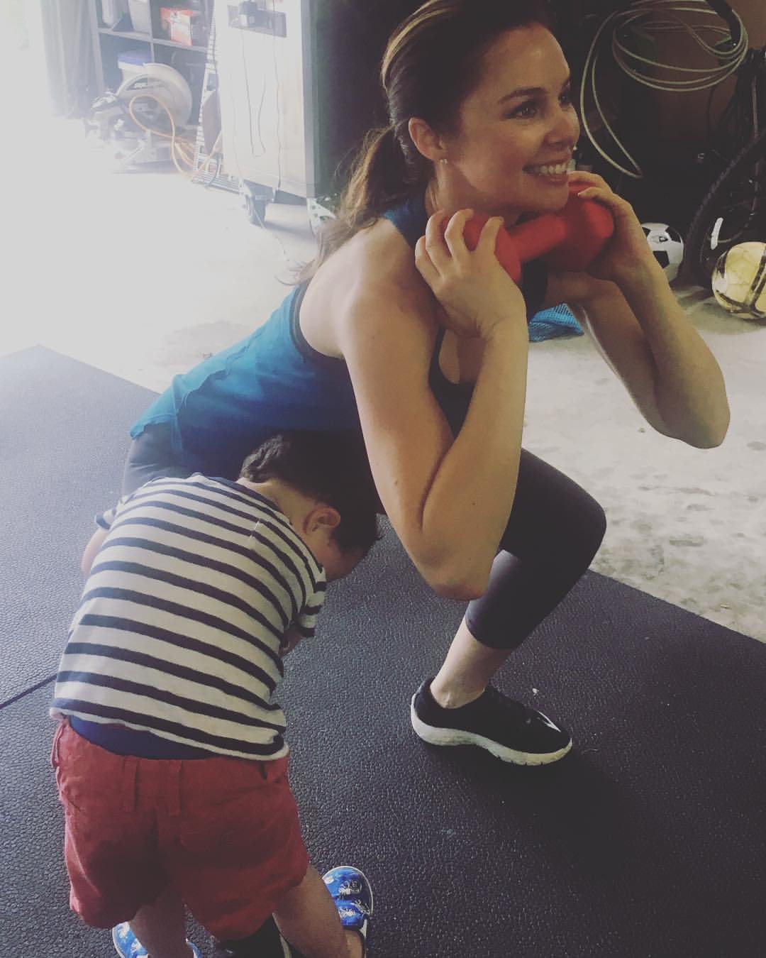 Testimony Tuesday - Meet Rainie Hutchinson, woman with brown ponytail working out in a blue tank top, she is front squatting a dumbbell with her toddler son wrapped around her leg